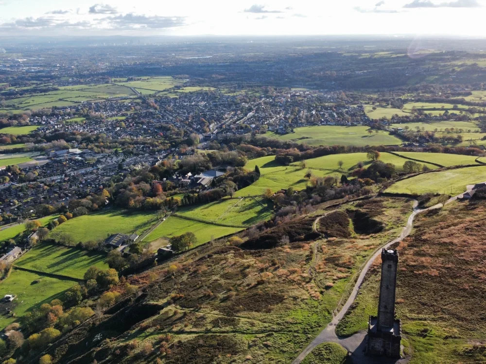 A photo taken from the hills around Bury, with the town in the distance and Peel Tower in the bottom right of the foreground.